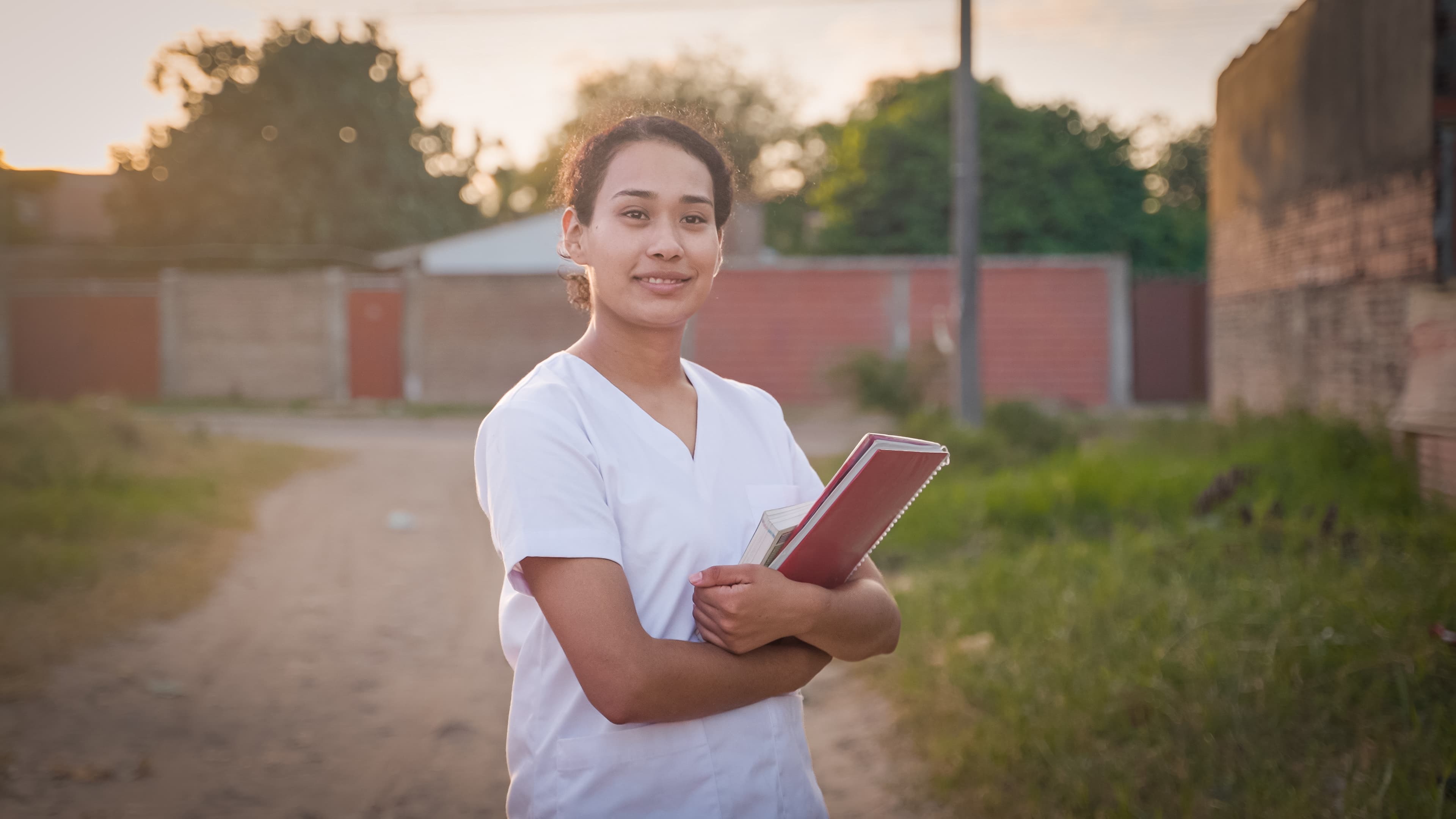 A young woman wearing white scrubs holds a book in her arms and smiles.