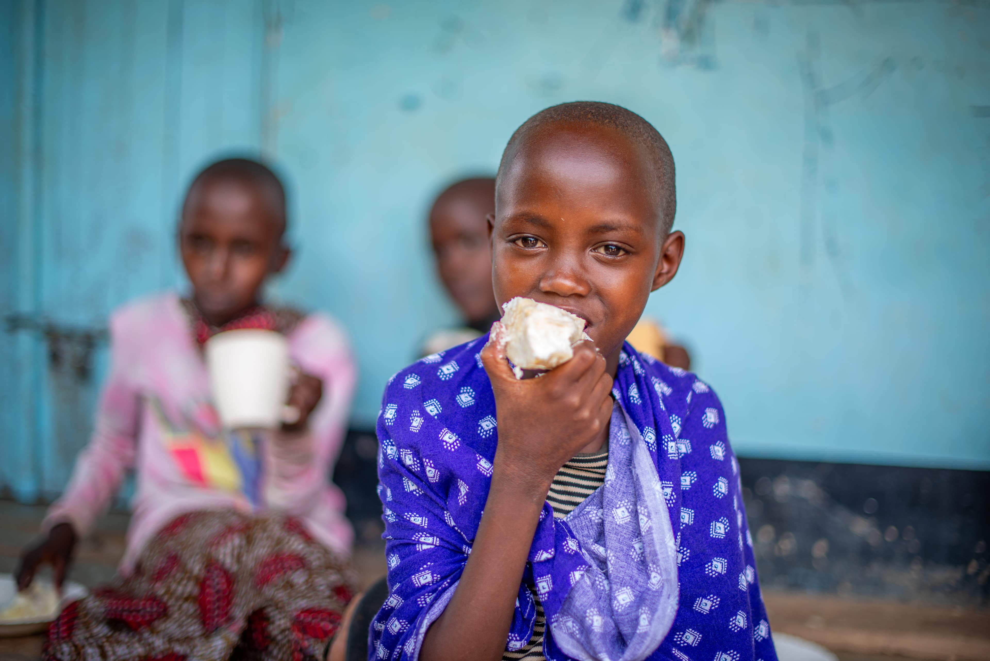 An African girl wearing a blue patterned shirt takes a bite of a cassava.