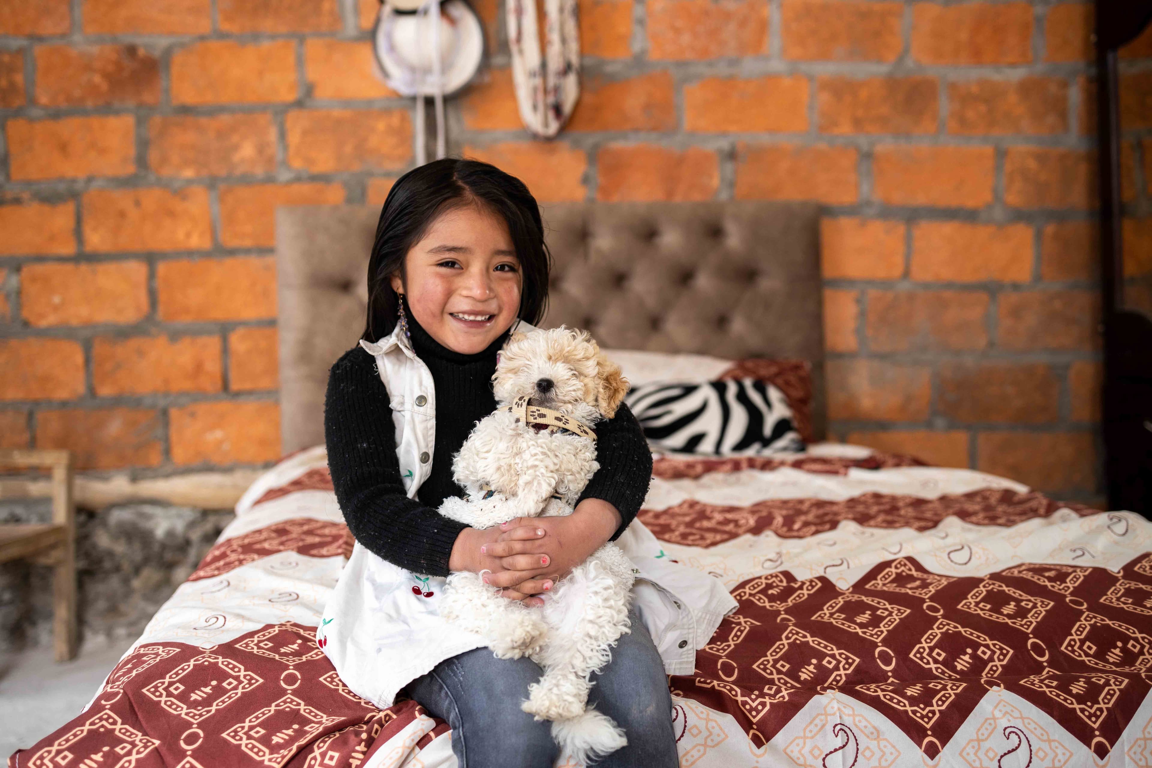 Young girl sits on a bed and smiles for the camera while holding a white puppy.