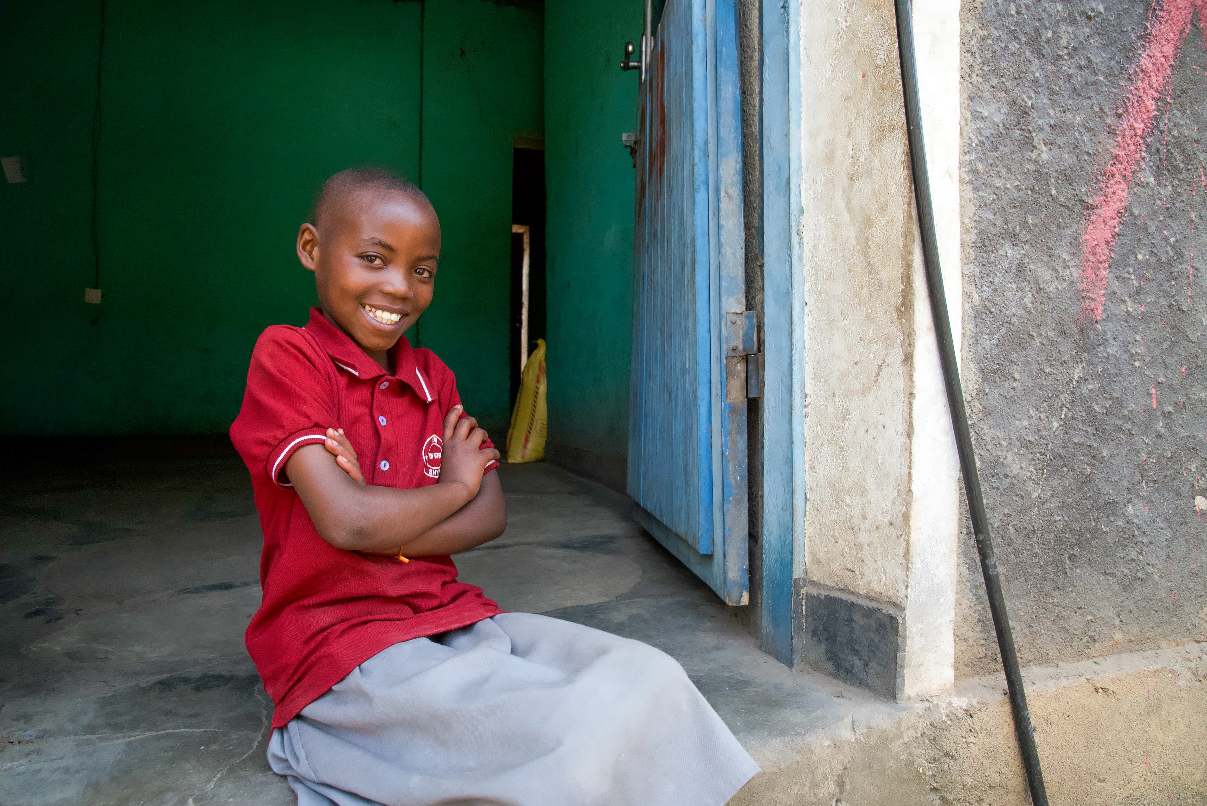 A young African girl wearing a red shirt sits in a doorway with her arms crossed while smiling.