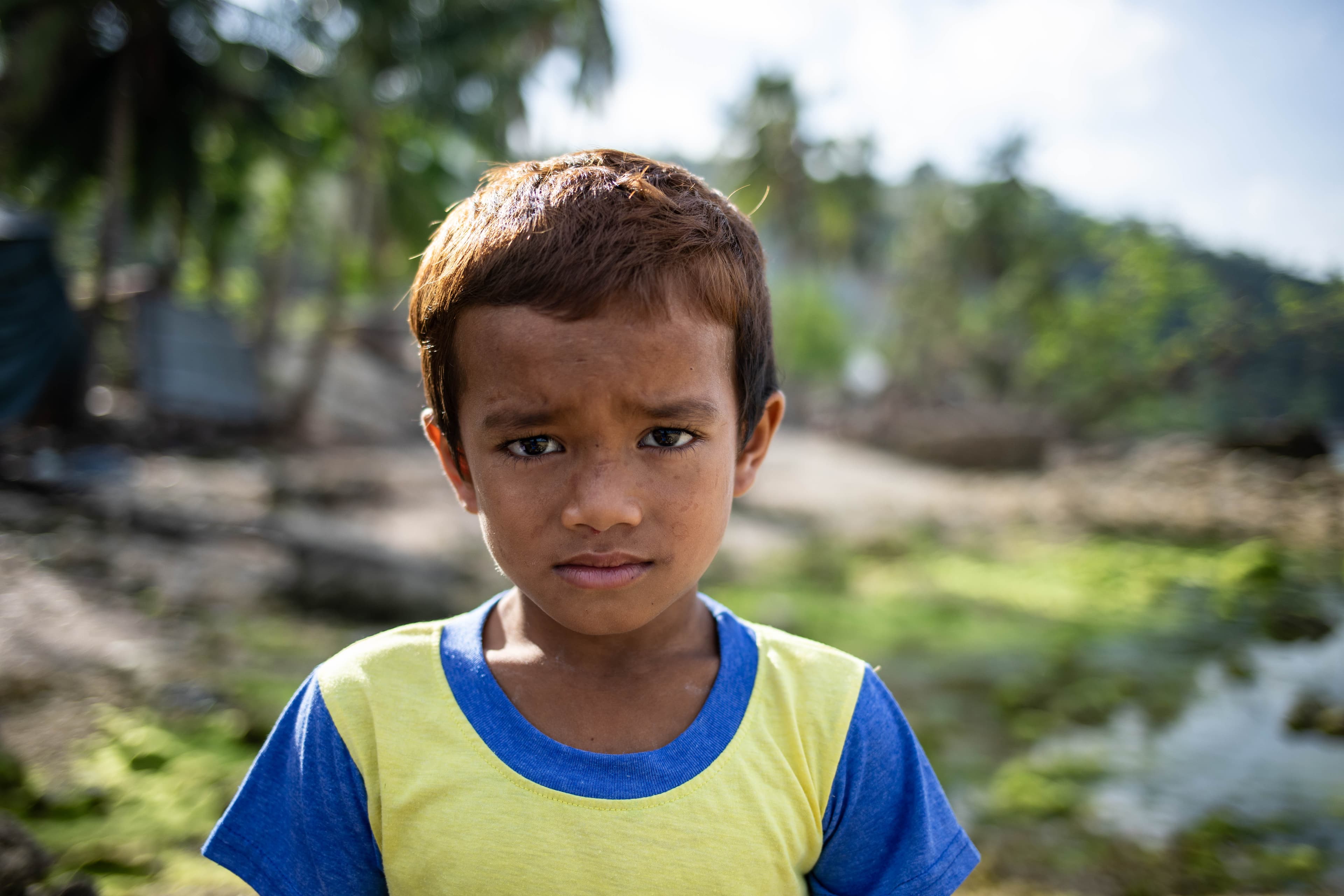 A young, Filipino boy wearing a yellow and blue T-shirt looks solemnly at the camera.