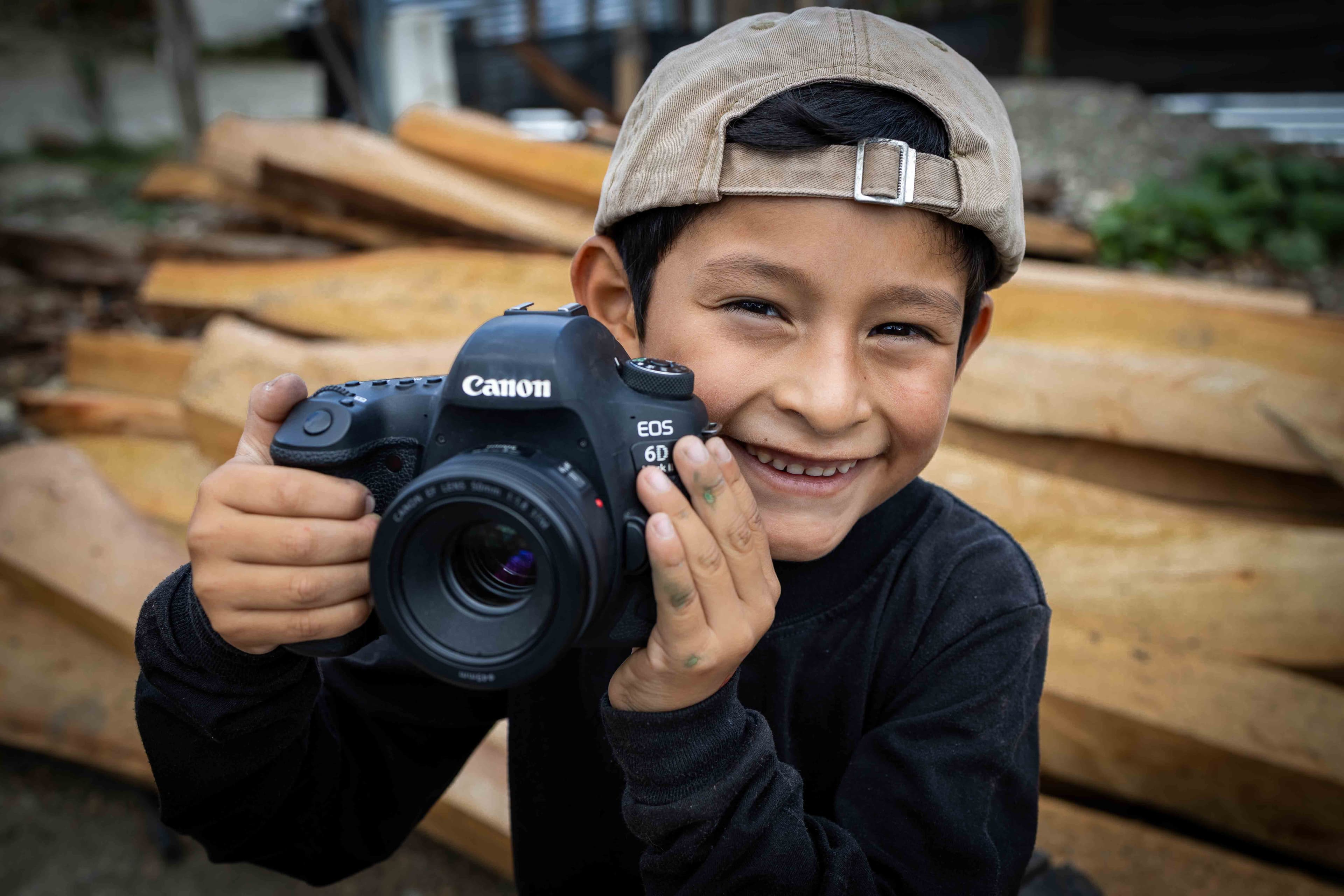 A young boy wearing a hat smiles for the camera while holding a camera next to his face.