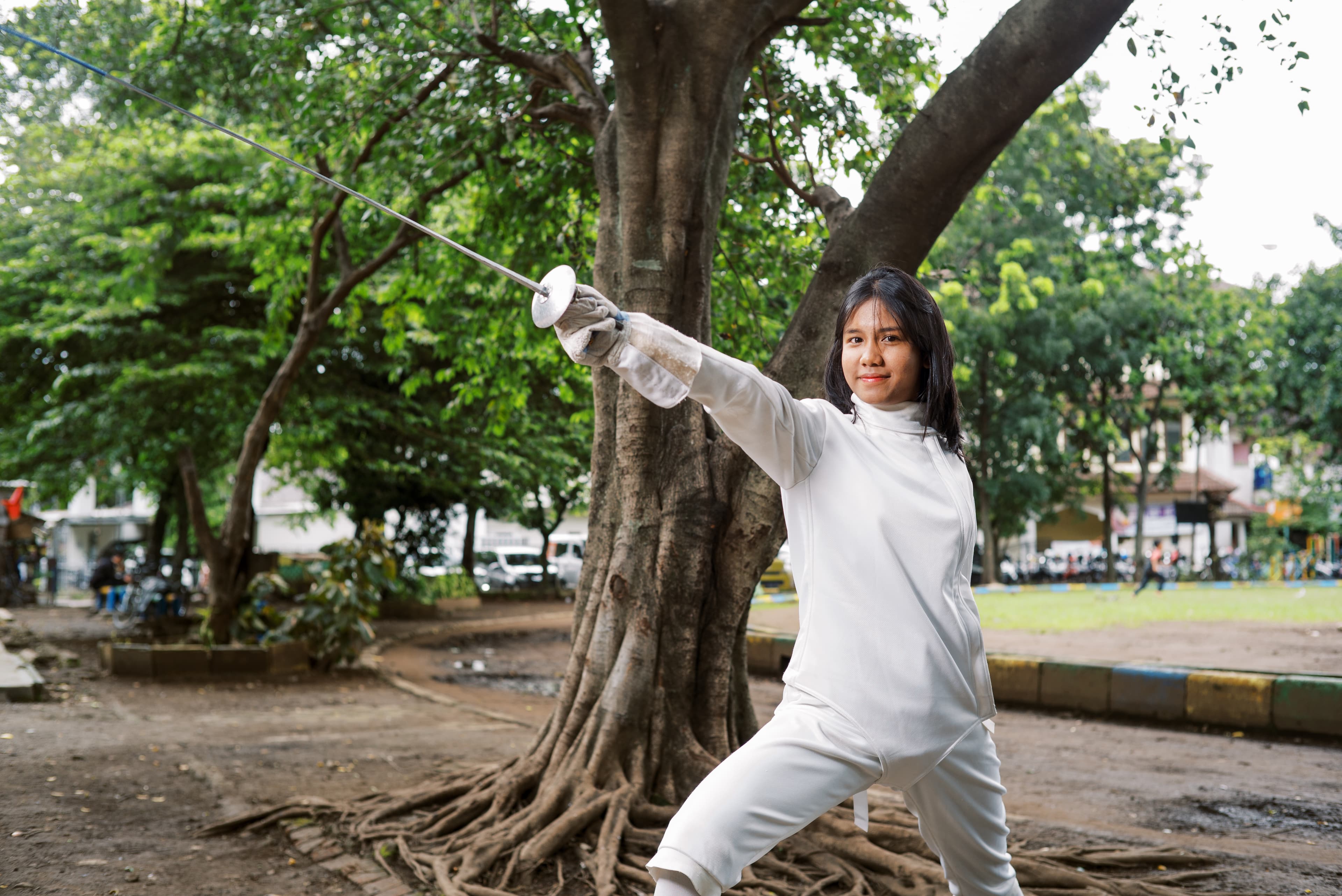 Young girl wears a white fencing uniform while holding her foil in the air and smiling.