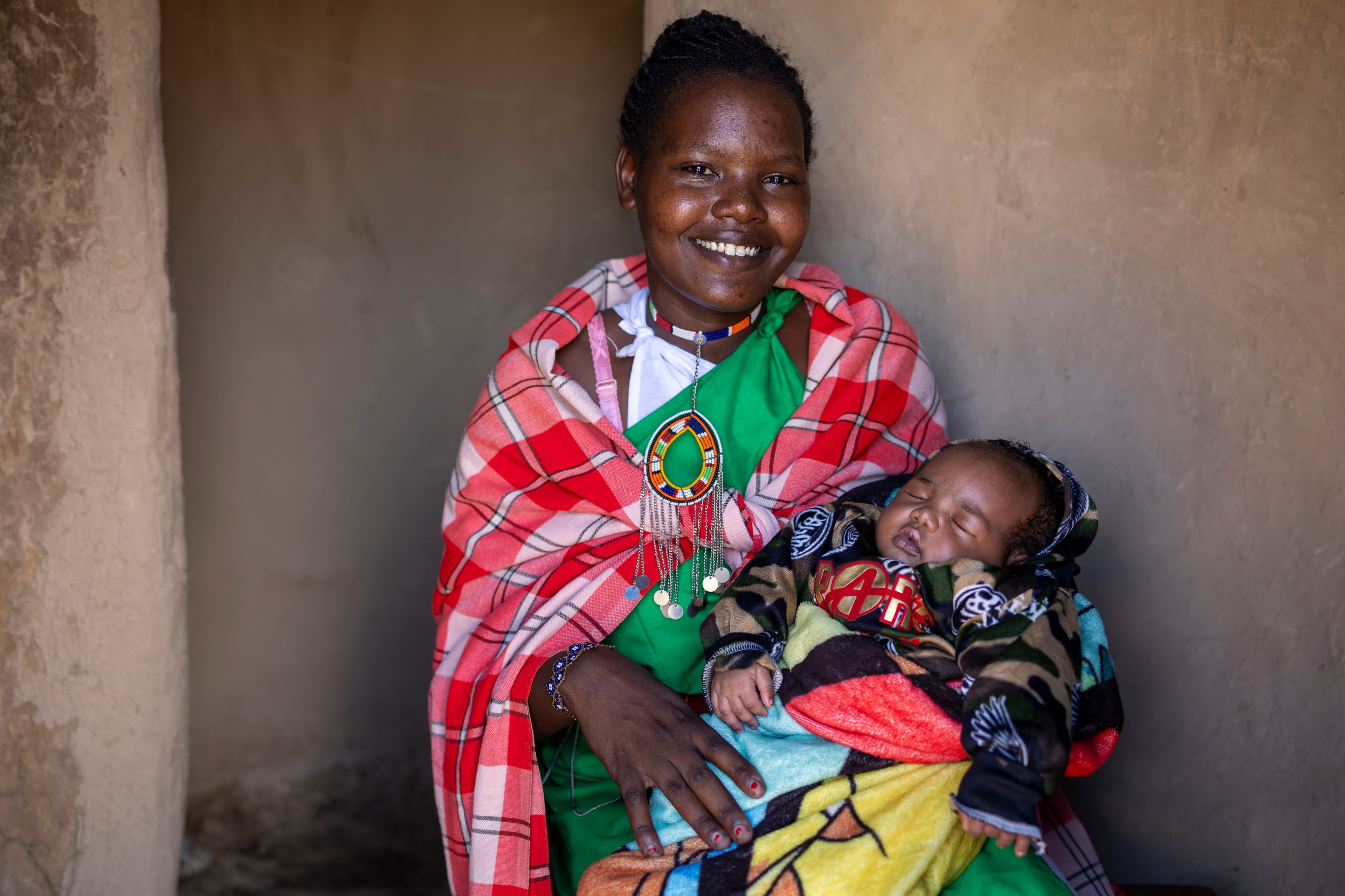 An African mom holds her sleeping infant baby while wearing a green dress and red, black and white shawl.