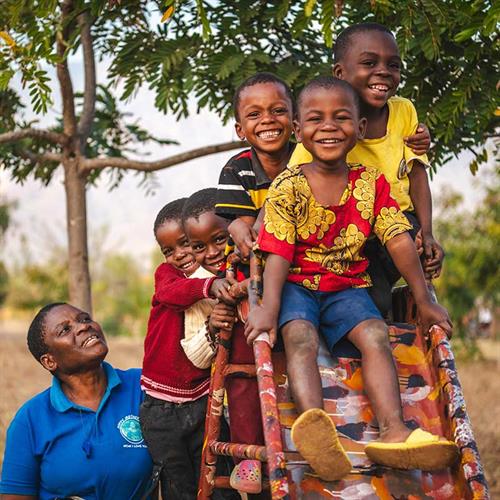 Pastor Beauty Senjele stands with some of the Program Participants at a playground who are climbing the slide, smiling together.