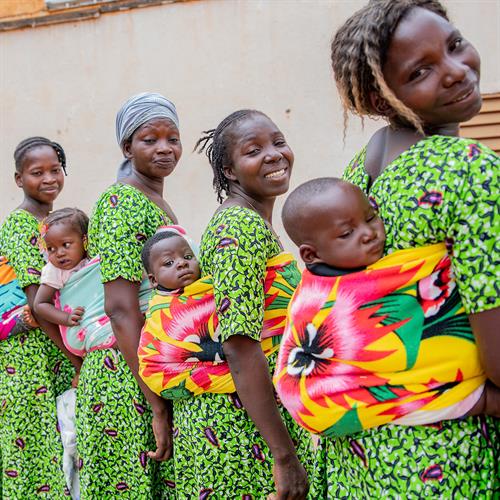 A group of moms smile at the camera while carrying their babies in traditional baby wraps