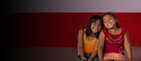 Two girls sitting close togehter in front of a red and white wall.