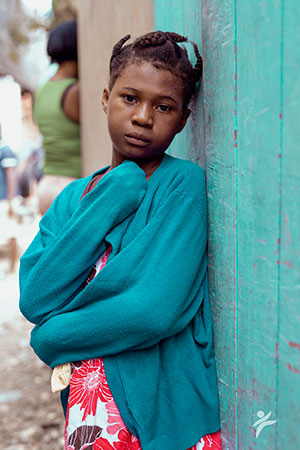 A solemn looking girl wearing a blue sweater leans against a wall