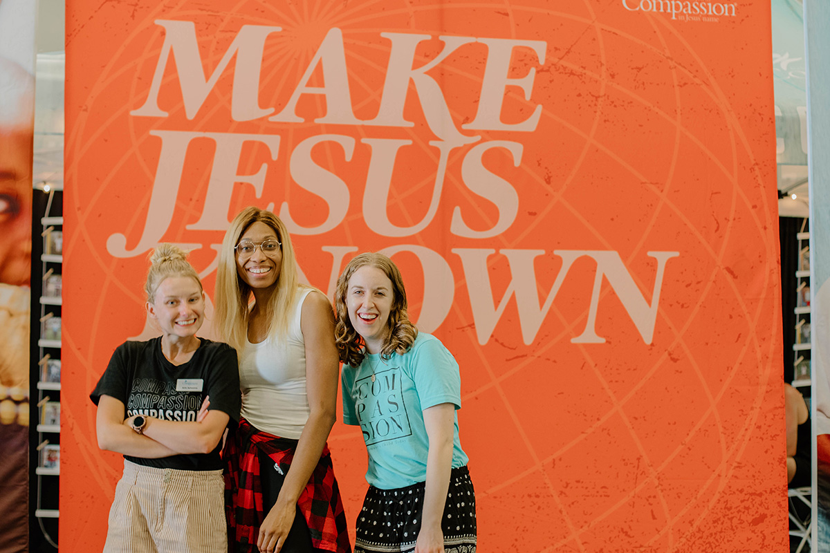 Erin and 2 other women stand in front of a "Make Jesus Known" banner