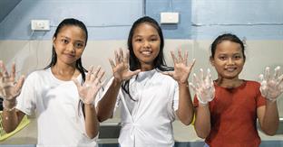 Standing in a bathroom area, three girls hold up soapy hands and smile at the camera.