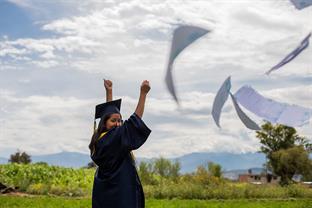A graduate celebrates in an open field by throwing papers in the air.