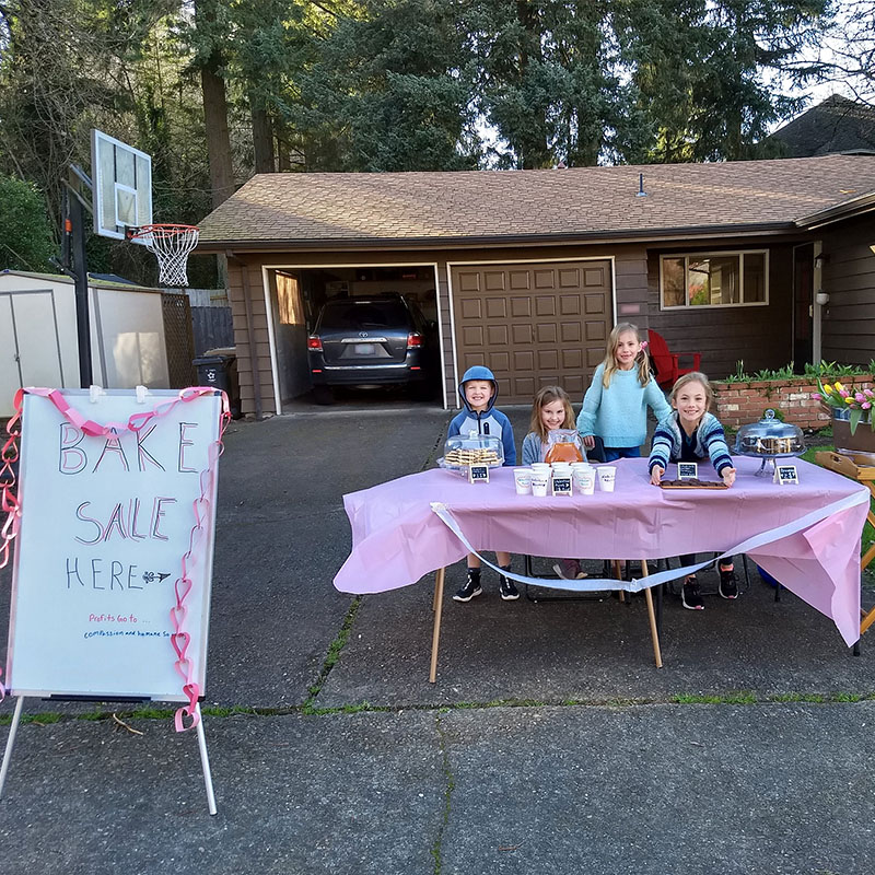 A group of children sit at a table outside of a house where they are hosting a bake sale.