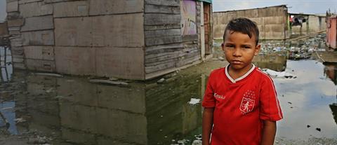 A solemn looking boy in a red shirt stands in front of a flooded house
