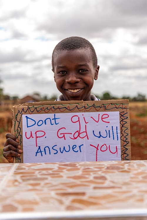 Smiling boy holding a sign that reads Don't give up. God will answer you.