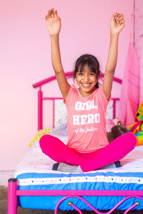 Young girl sitting cross-legged on a bed. She is smiling and raising her hand in the air.