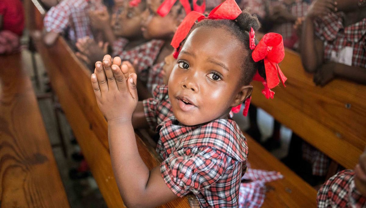 Little girl with red bows in her hair kneeling on a bench with her hands together in a praying pose.