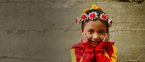 A smiling girl with a flower wreath headband places her hands on her cheeks
