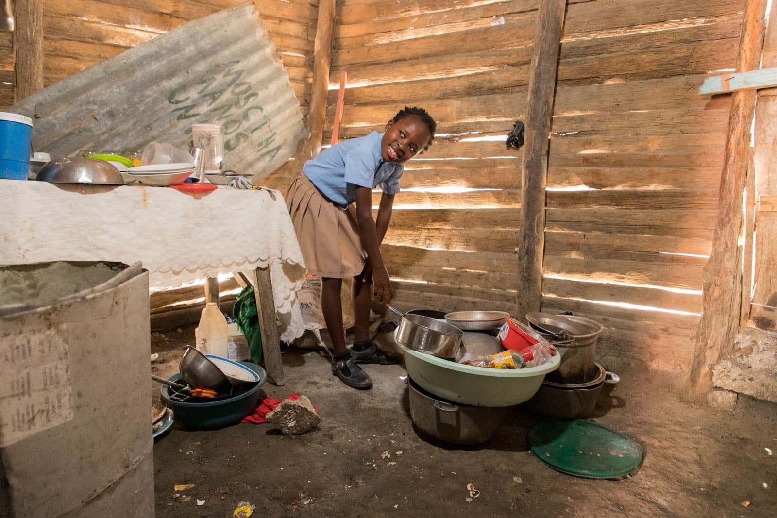 Yosaira Yaque, 9 years old, is wearing her school uniform, a khaki skirt, sky blue shirt, black shoes, and black socks. She is cooking in the kitchen of her home, which is in the left part of her and her sister’s room. The walls of her house have holes in them which allow water to get in when it rains.