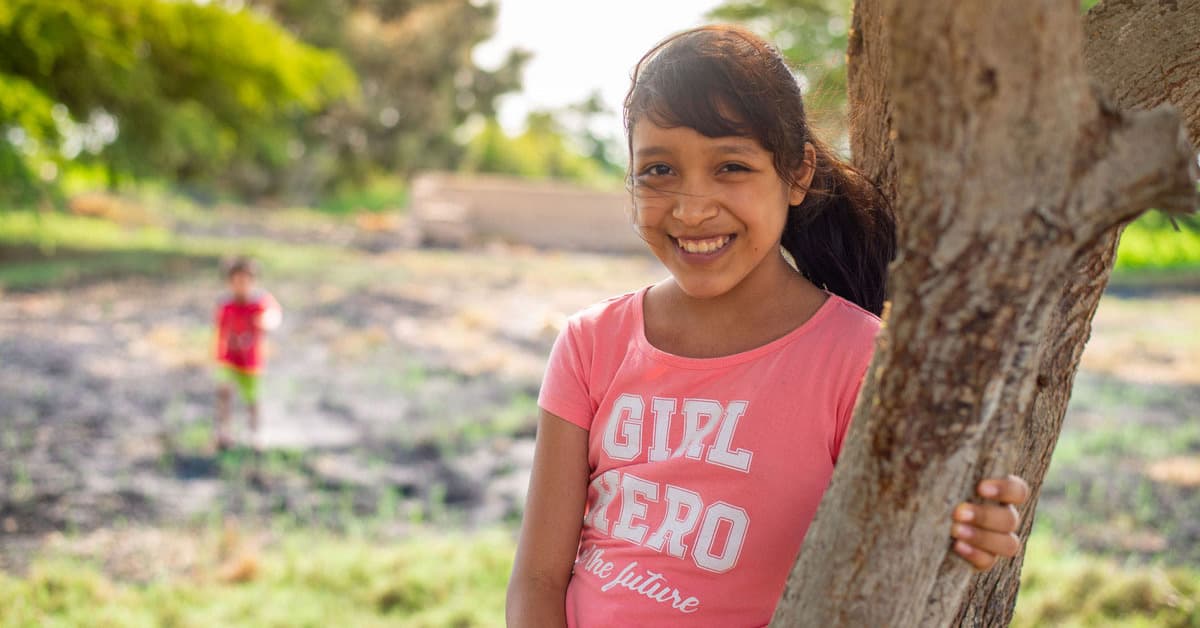 Girl in a pink shirt leaning against a tree smiling.