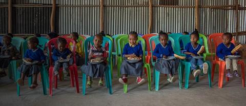 A group of Ethiopian children sit in plastic chairs while holding plates of food in their laps