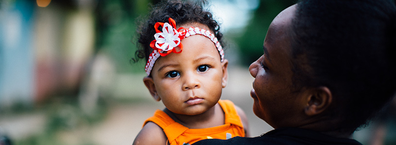 A Dominican baby in orange clothes looks over her mother's shoulder.