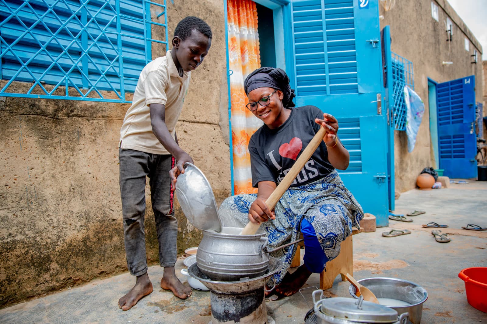 Adèle is wearing a black shirt that says, "I love Jesus." She is sitting down outside her home and is cooking. Her younger brother, Abdoul, wearing gray pants and a tan shirt, is helping her by pouring corn flour into her pot. Their home is behind them. It is tan with blue doors and shutters, and there is an orange and white curtain hanging in the doorway.