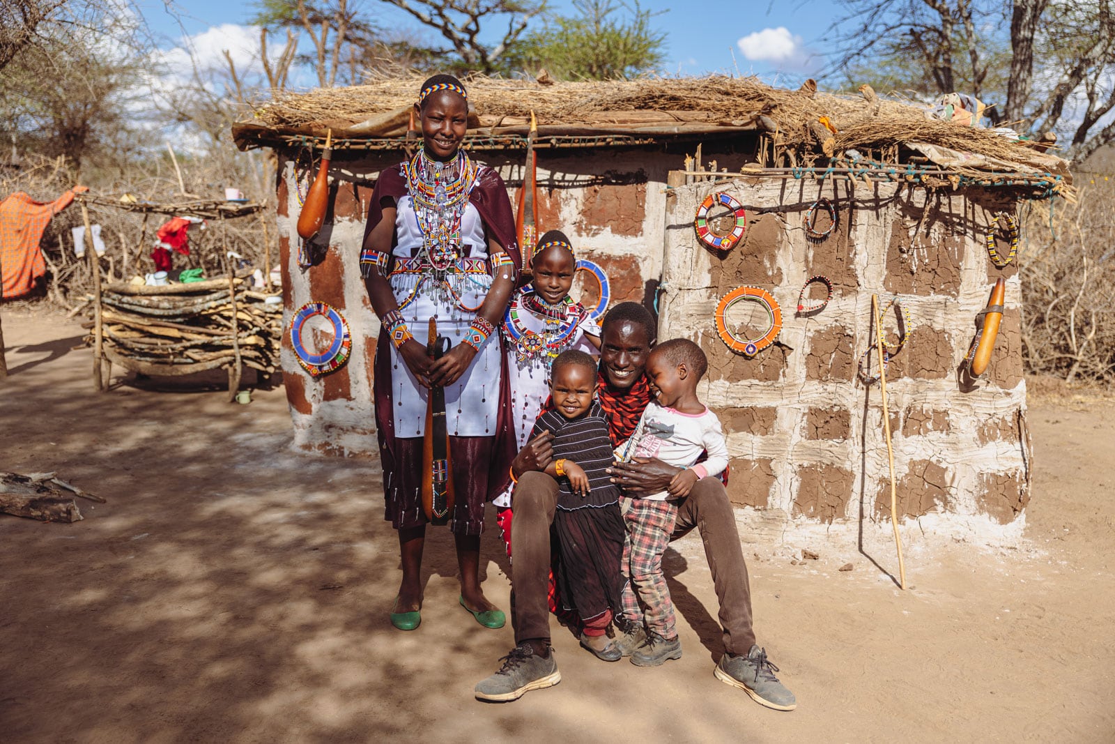 Pesi, her mother, father, and her siblings Enock and Tasha are sitting outside their home. Behind them a wall decorated with beaded jewelry. Pesi and her mother are wearing traditional clothing