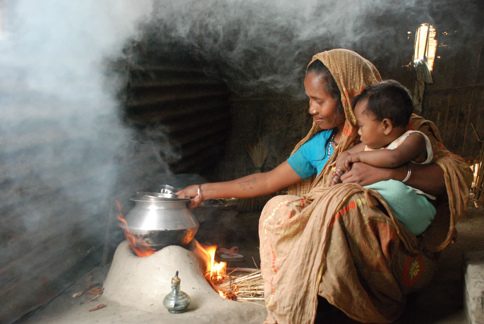 Woman sitting by a mud fire pit. She is holding her child in her arms, looking down as she holds a silver metal cooking pot over an open fire smoking stove cooking.