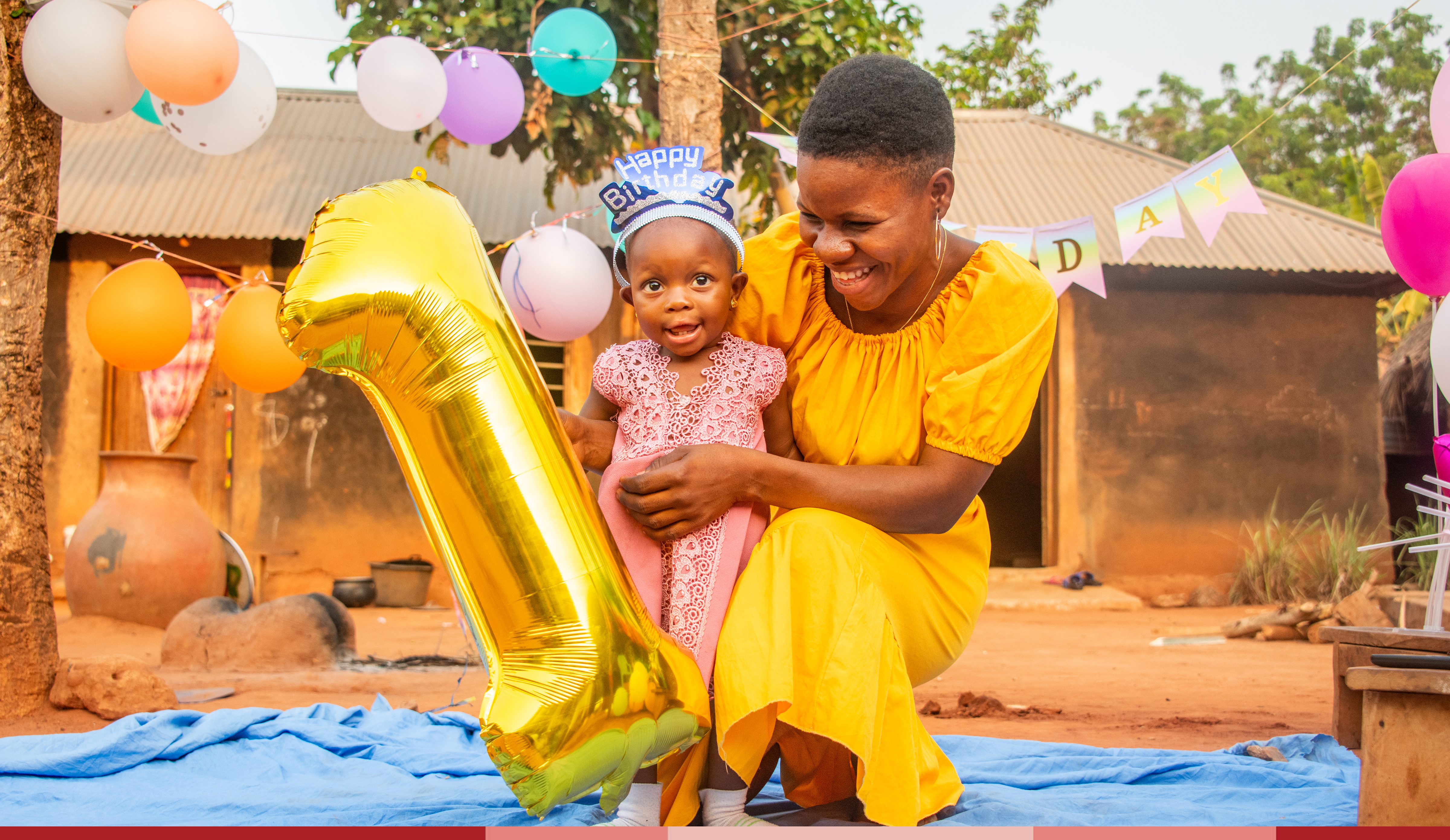 Davi is wearing a yellow dress while holding Yawa and a one-shaped balloon at Yawa's birthday party