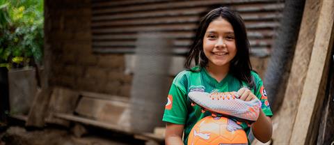 a girl holds her soccer cleats and soccer ball