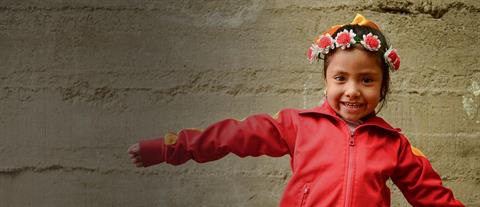 A young girl in a red jacket and a white flower crown stands in front of a cement wall with her arms spread out 