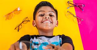 Young boy lies on a yellow and pink background and holds up glasses to the camera. He smiles brightly.