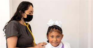 A young girl wearing a white bow smiles for the camera as a female doctor uses a stethoscope to listen to her breathing.