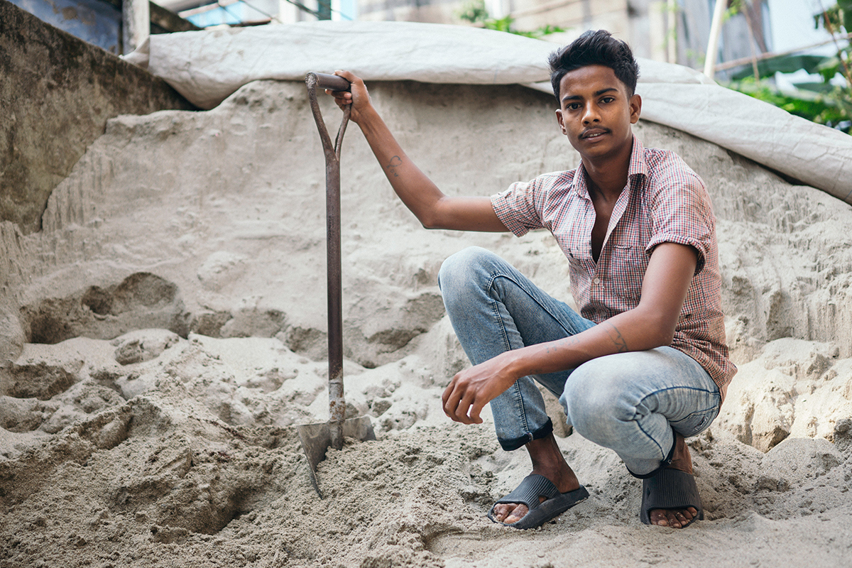 Shanto sits in front of a sandpile holding a spade