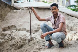 Shanto sits in front of a sand pile holding a spade