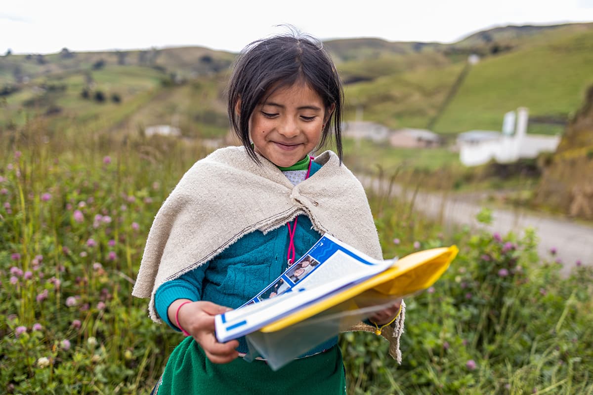 Girl wearing traditional clothing: a blue sweater, white shawl, and a green skirt. She is standing in a field of wild flowers and is reading a letter from her sponsor.