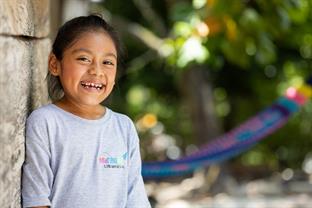 Young girl leans against a brick wall and smiles brightly at the camera.