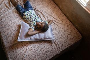Young boy lying on a bed with his hands behind his head looking up and smiling at the camera.