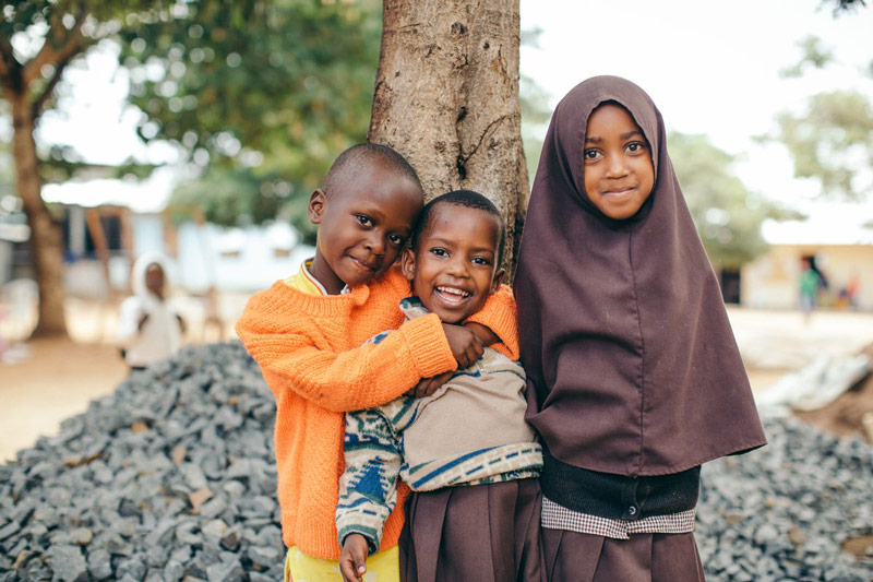 Three children smile and stand in front of a tree