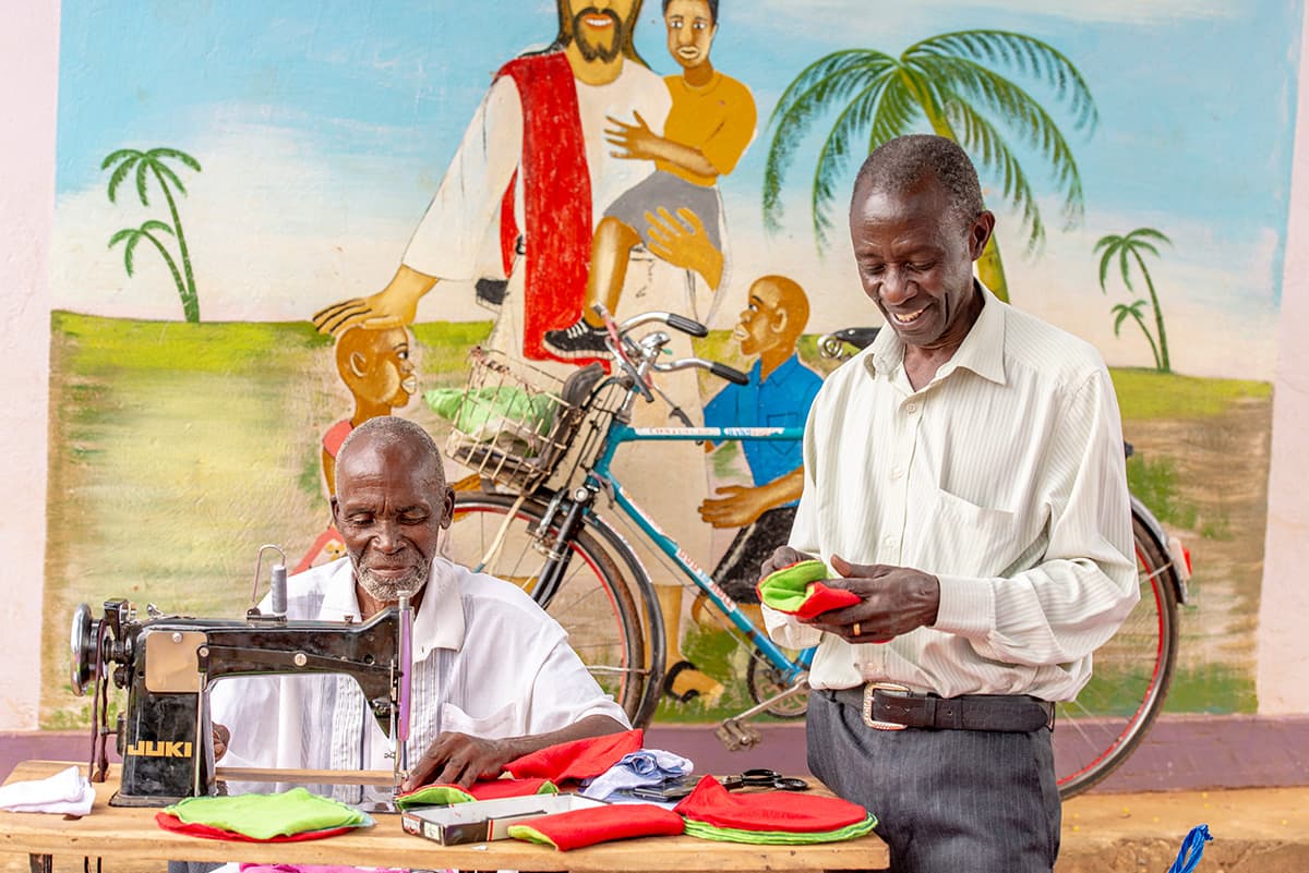 Pictured here are two men working together to sew reusable sanitary towels. They are smiling as they work. There is a picture of Jesus and a bicycle in the background.