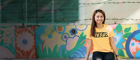 A young woman sits on a floral painted wall and smiles with a yellow "Jesus" shirt on