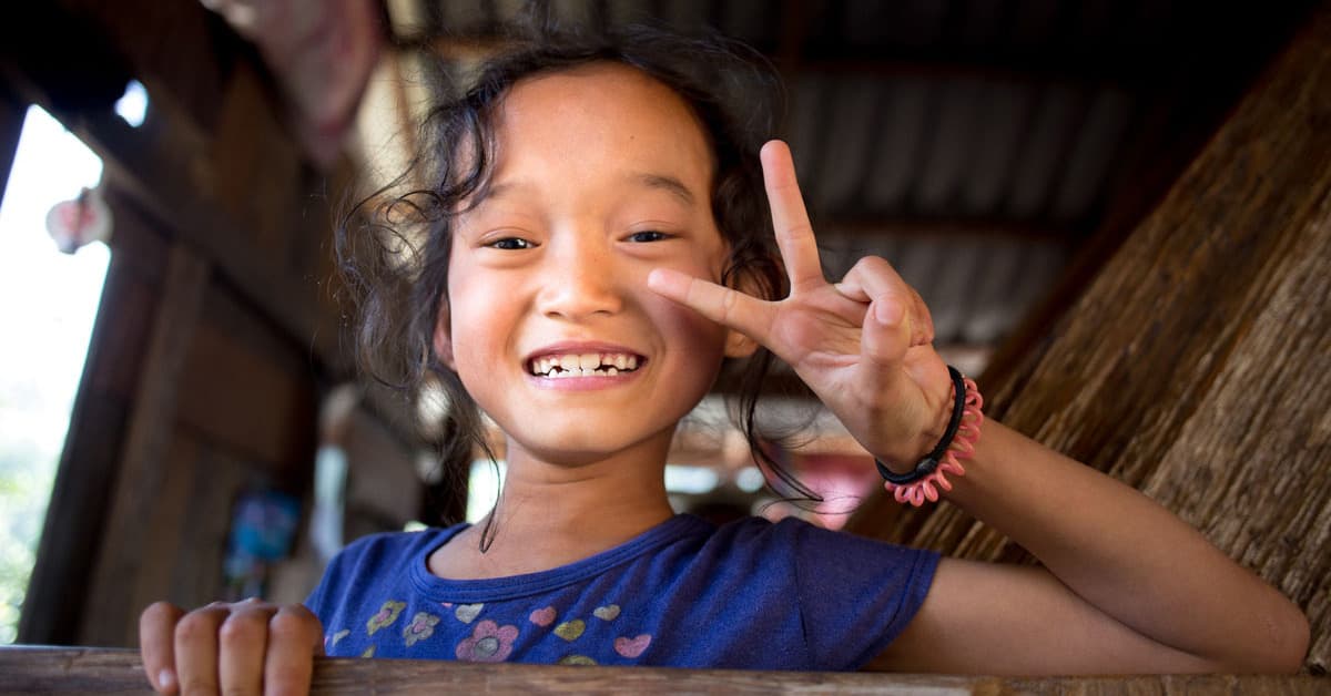 A smiling little girl with the fingers of her left hand making the peace sign.