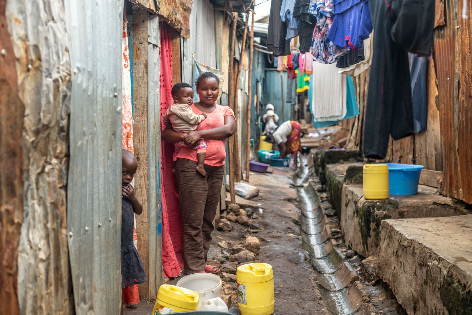 Wanja is standing outside in the alley of the slum where she lives. She is wearing a red shirt and brown pants. She is holding her youngest child. There are clothes hanging up on clotheslines behind her.