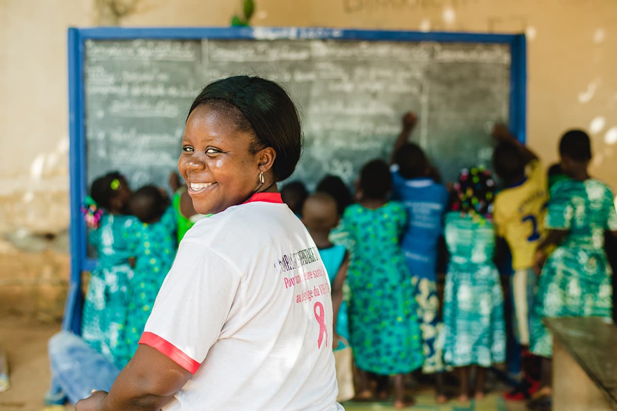 Compassion staff wearing a white shirt looks back over her shoulder sitting in a  classroom with a group of children lined up facing a chalk board writing lessons.