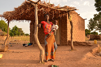 An African woman holding a baby stands outside of her mud home.