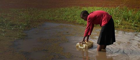 A woman in a red shirt bends at the waist to fill a jerry can with water from a pond
