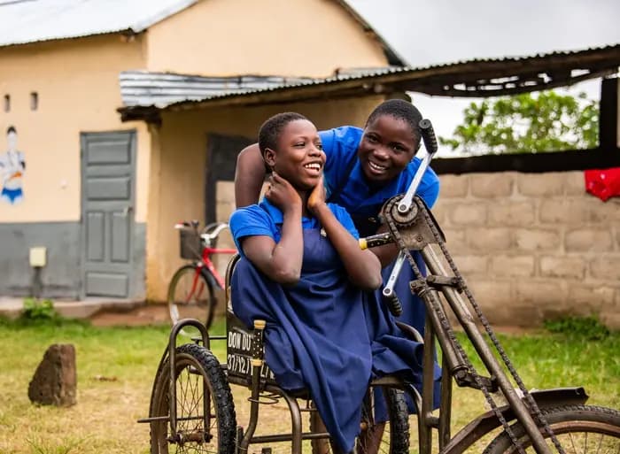 Mawussi is in the yard at the center with her friend Christina. Mawussi is sitting in her wheelchair and Christina is leaning over to talk with her. Both are wearing the center's uniform of a navy blue skirt and blue t- shirt.