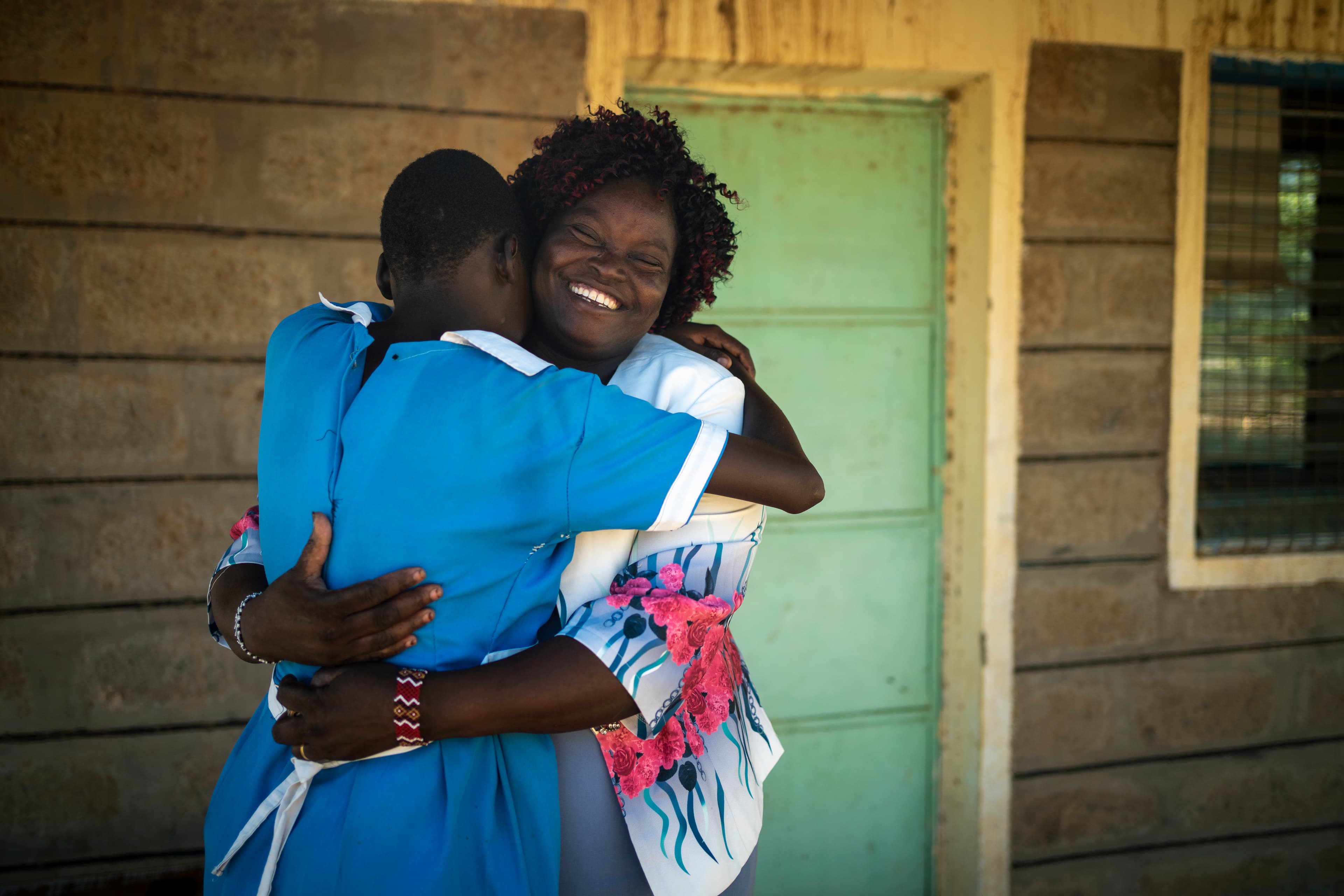 Two African women hug each other in front of a green door.