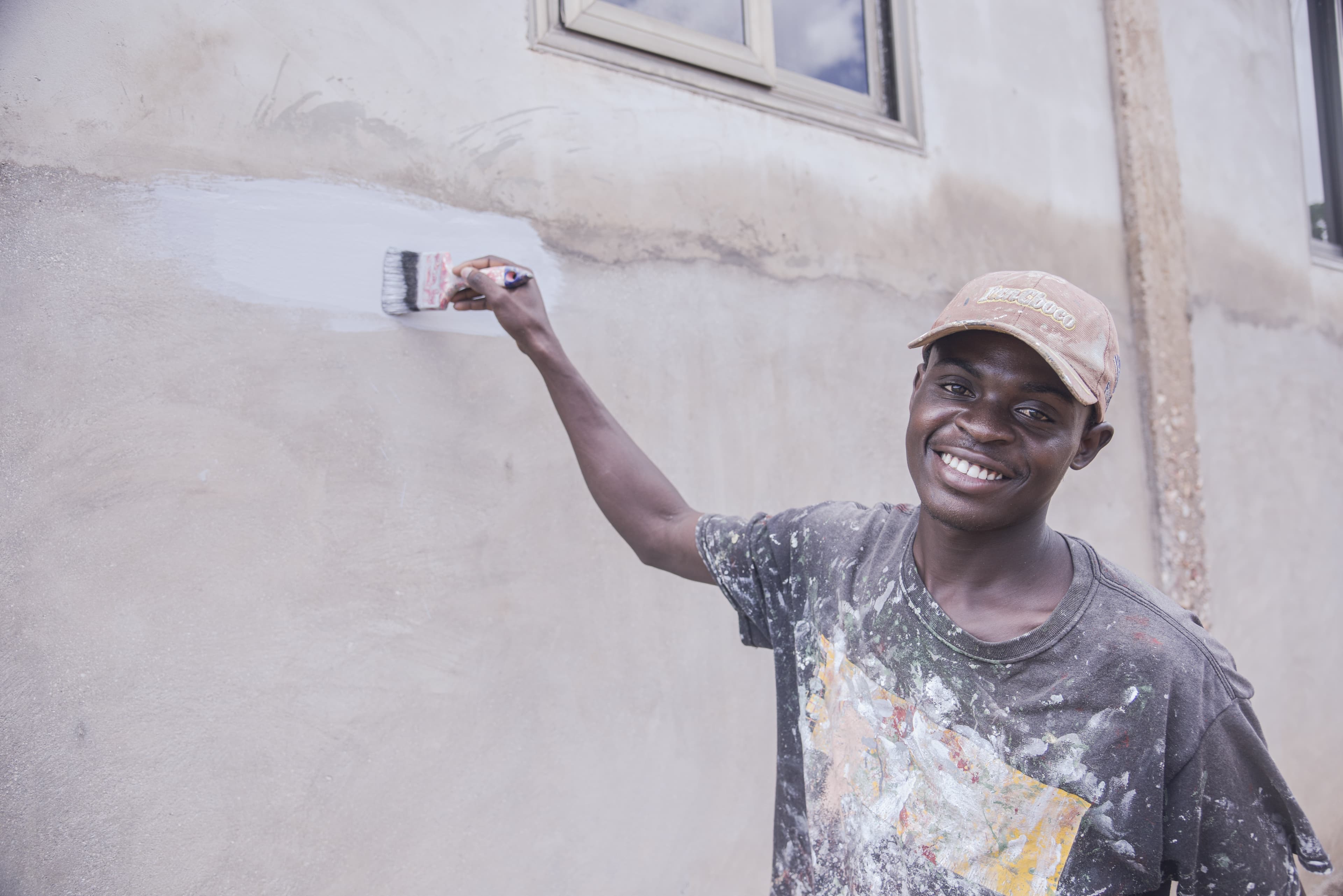 A man stands with a paintbrush in his hand looking at the camera as he paints the side of a building.