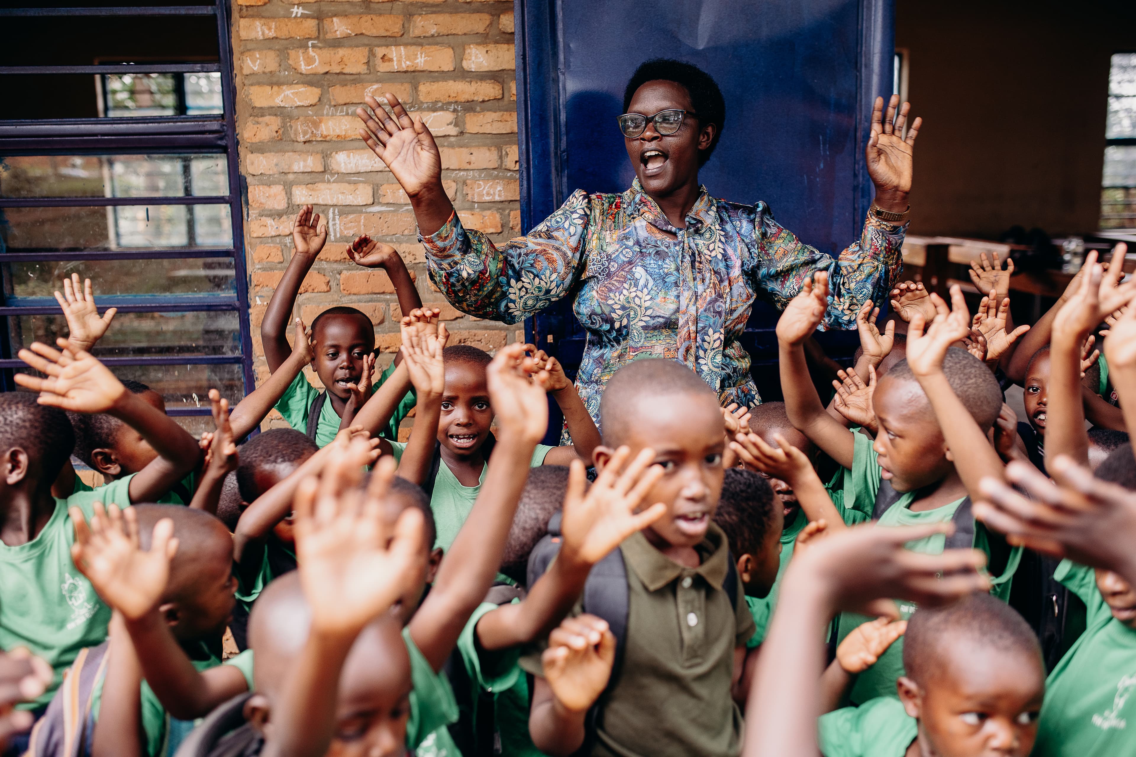 A group of African children wearing school uniforms raise their hands and dance in front of a building.